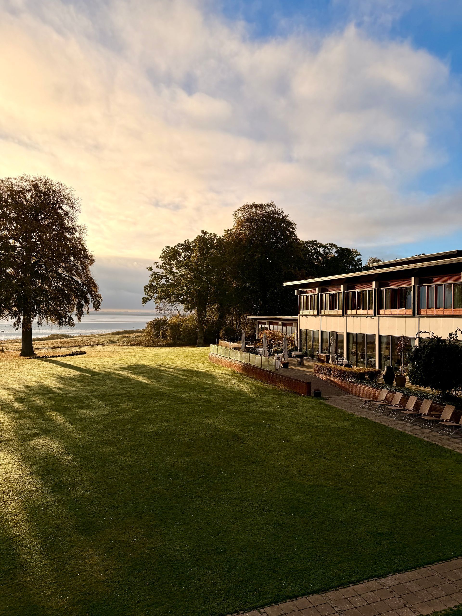 Ydersiden af ​​en lys bygning med udendørs siddepladser og et glasindkapslet område; overskyet himmel over hovedet.