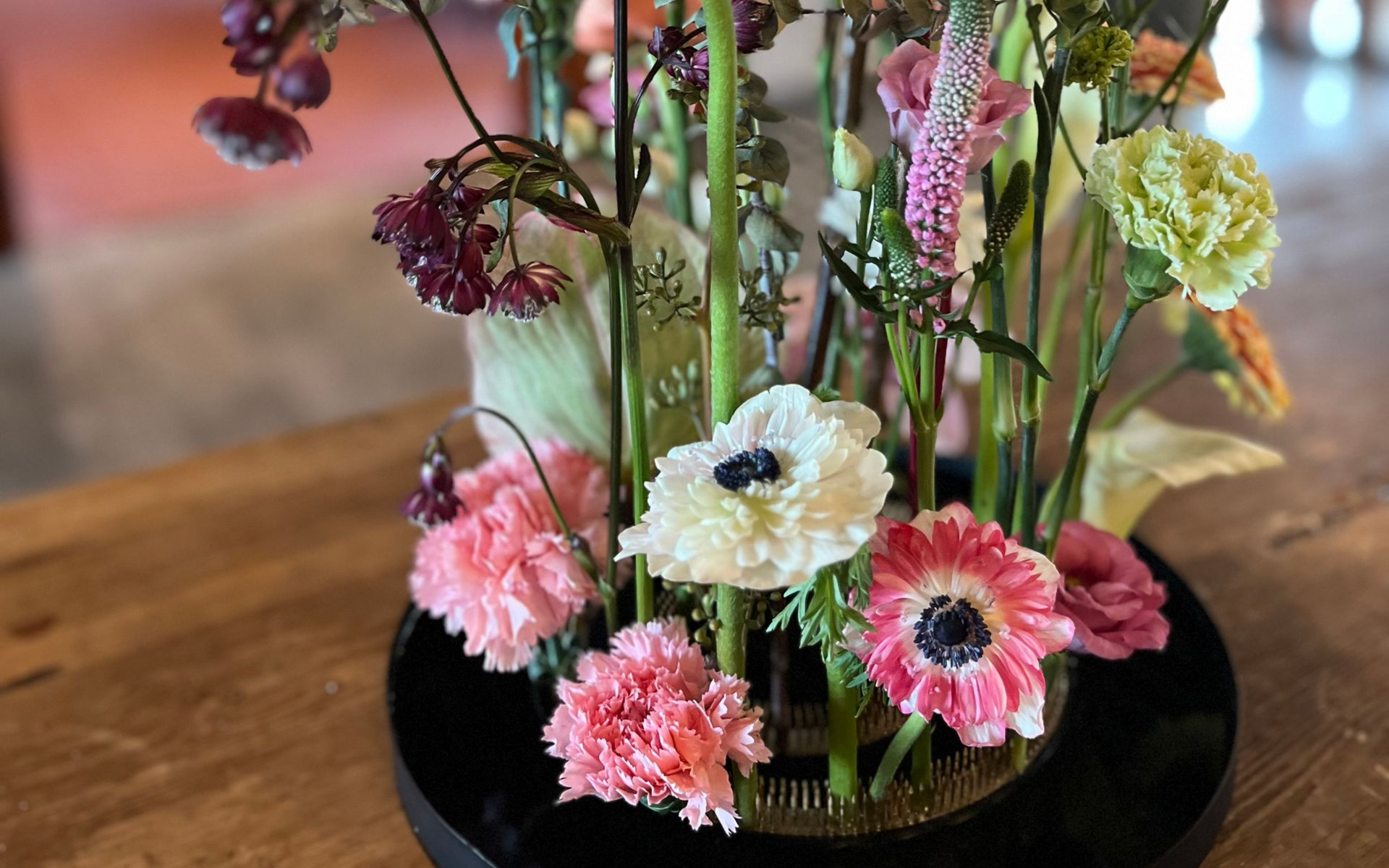 Blomsterarrangement med lyserøde og hvide blomster på et træbord, foran en sløret baggrund.