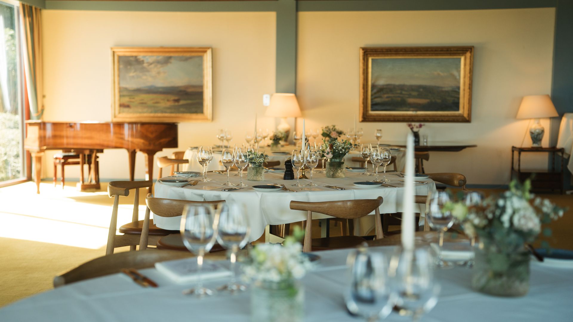 Elegant spisestue med et rundt bord dækket til et måltid; klaver og landskabsmalerier i baggrunden.