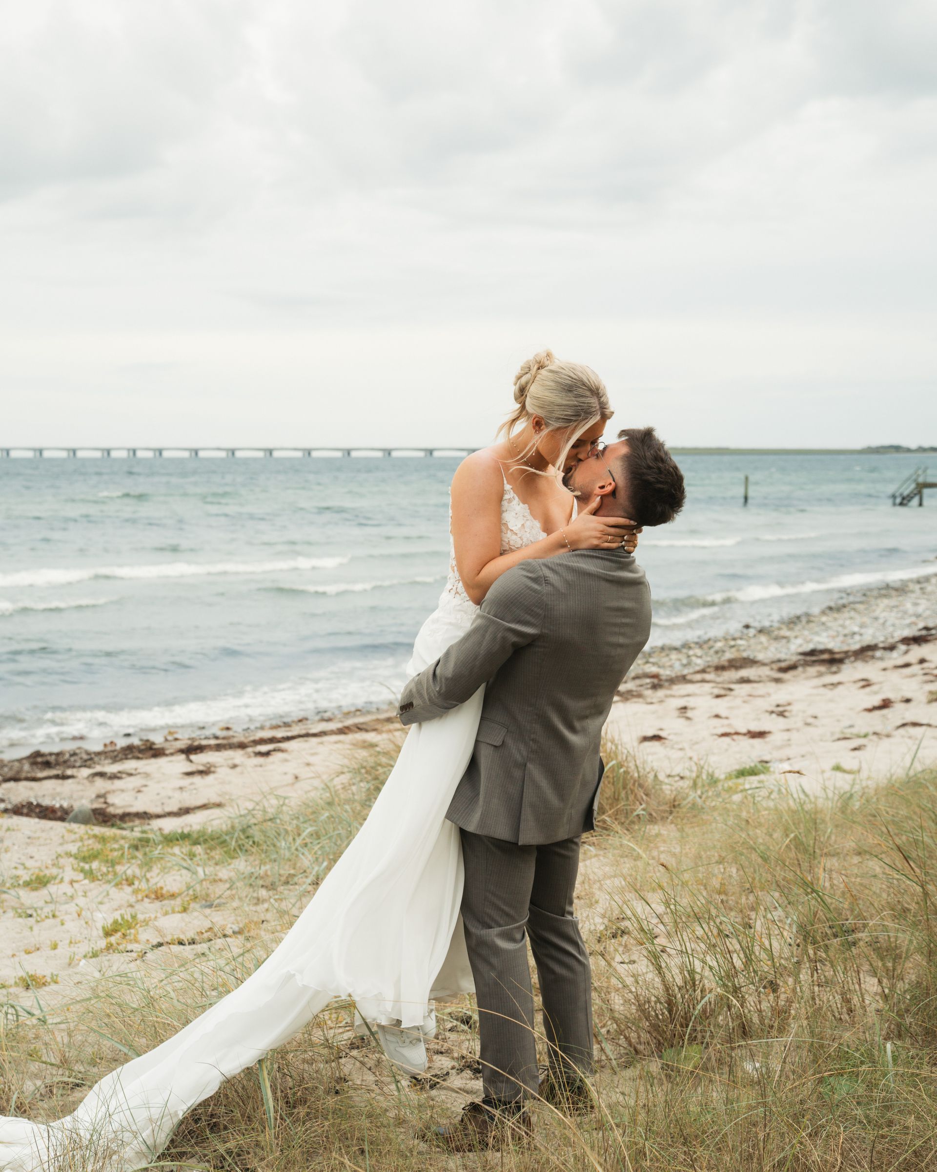 Mand løfter en kvinde i brudekjole på en strand, mens hun kysser. Hav og bro i baggrunden.