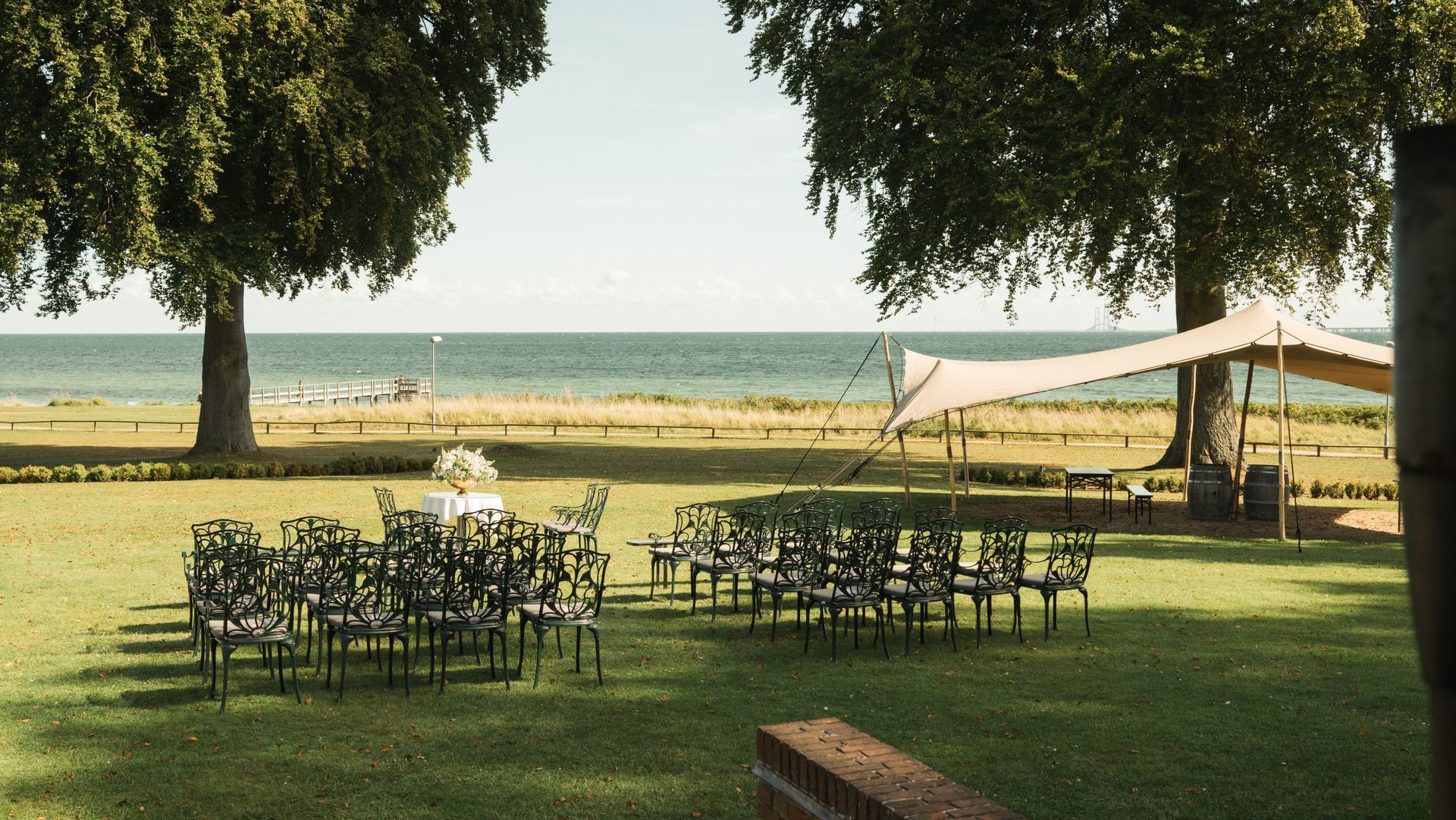 Græsplæne med stole opstillet til et arrangement, med udsigt over en strand og hav under et telt.