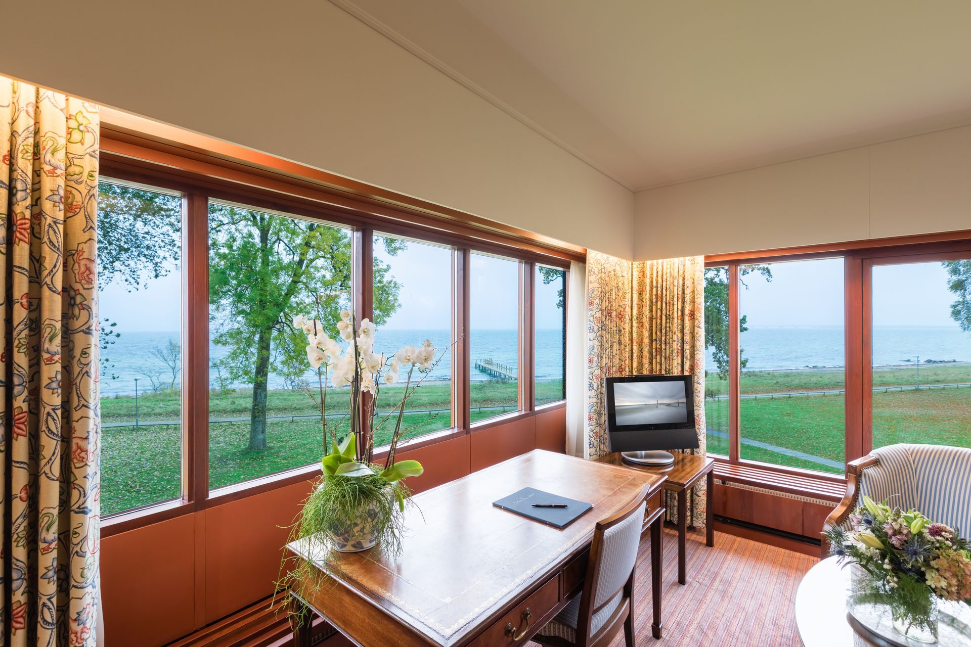 Skrivebord i værelse med store vinduer med udsigt over vand og grønt landskab. Fjernsyn og blomster.