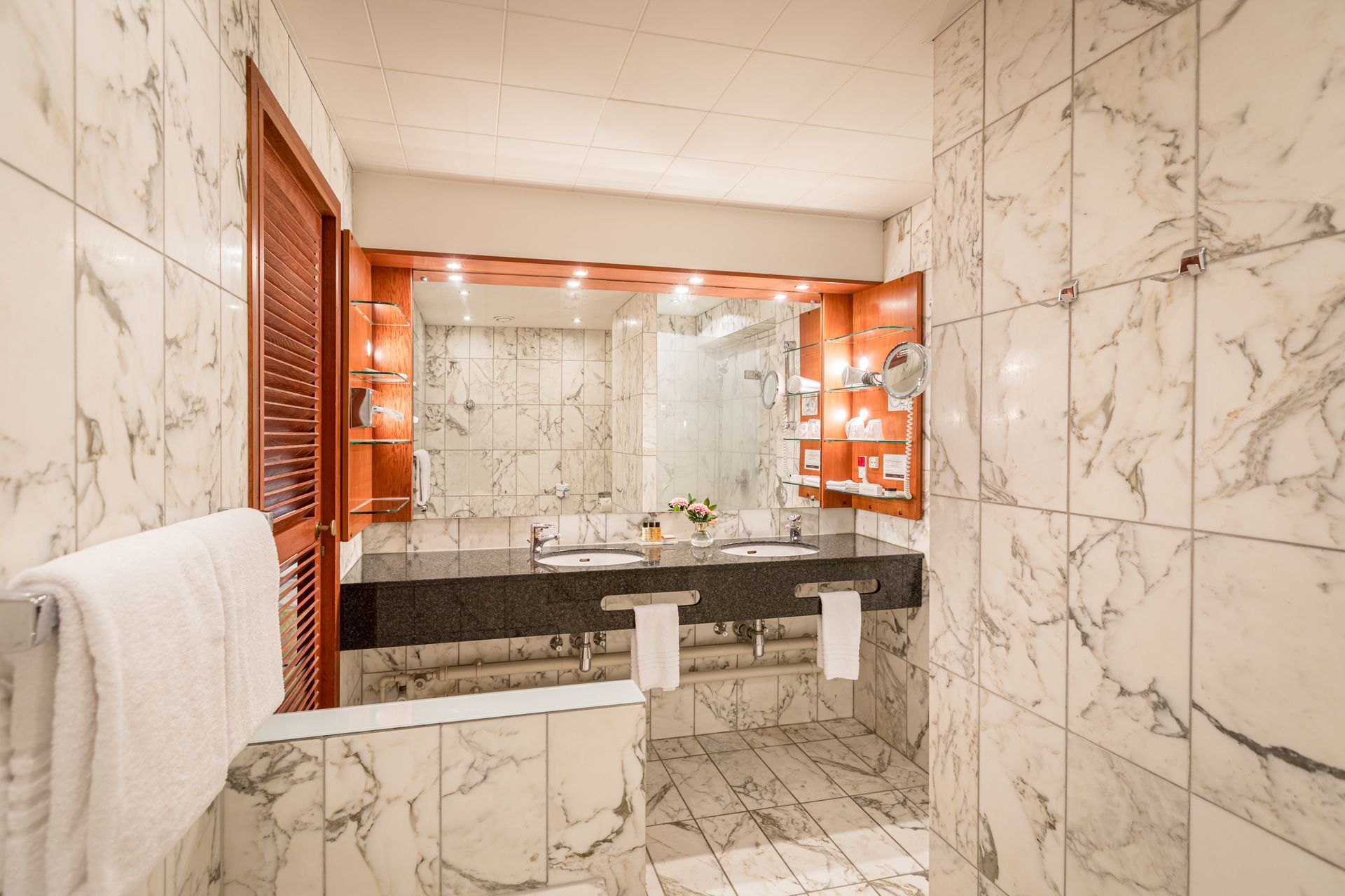 Elegant badeværelse med marmorfliser, dobbelt håndvask, stort spejl, trædetaljer og håndklædestativ.