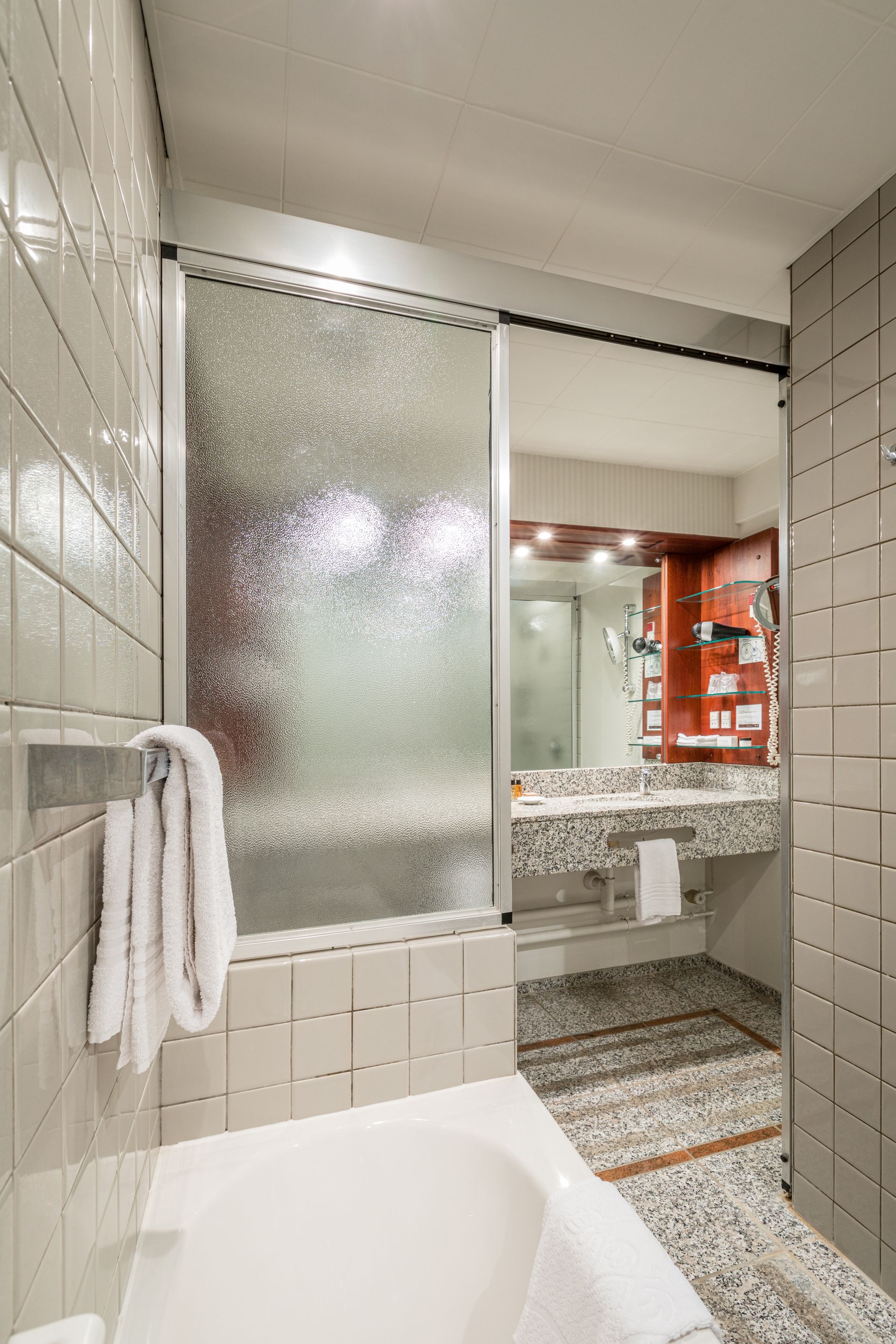 Badeværelse med badekar, skydedør i matteret glas, håndvask og flisevægge.