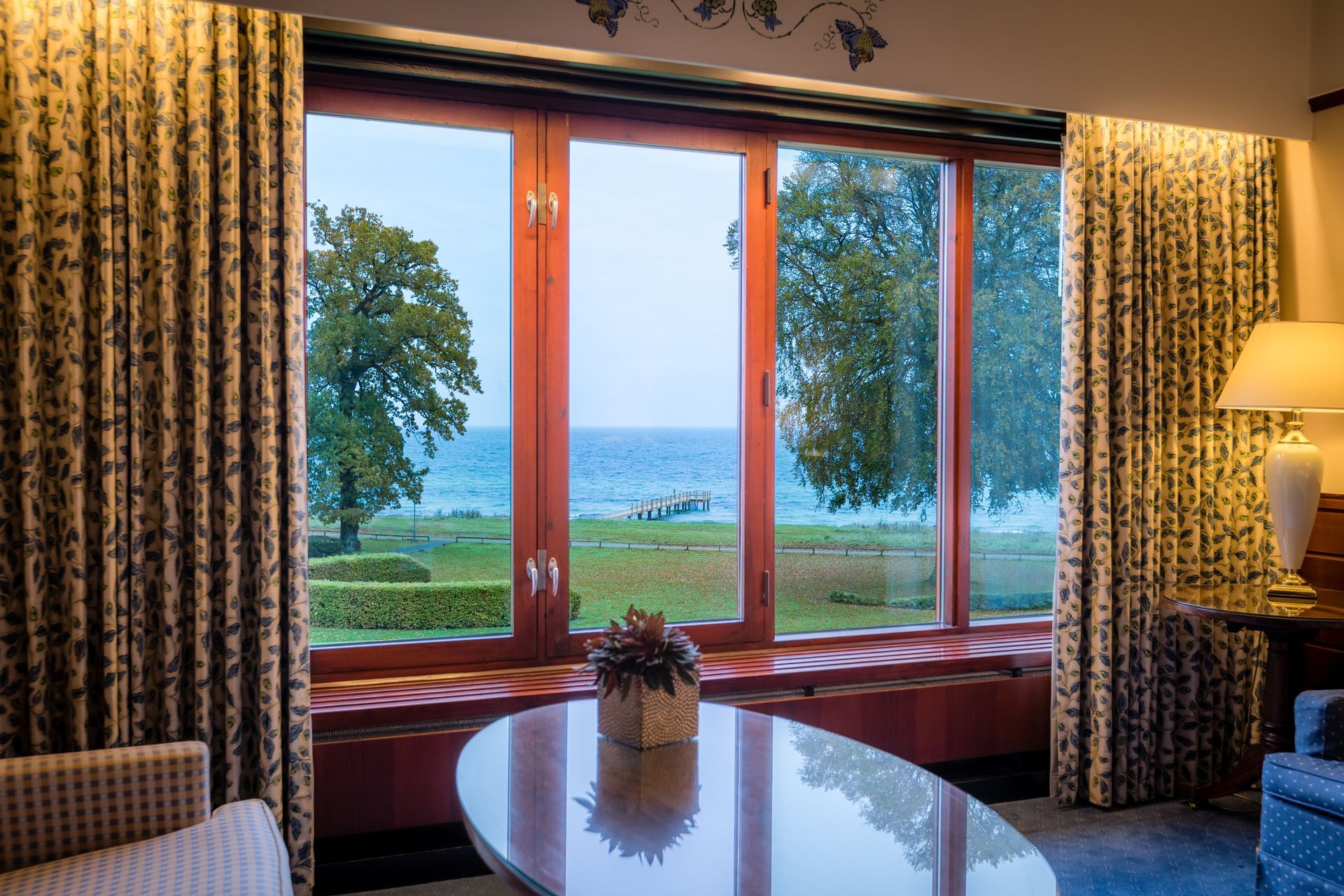 Indvendig udsigt over et vindue med udsigt over et vandområde og træer, med gardiner og et bord.