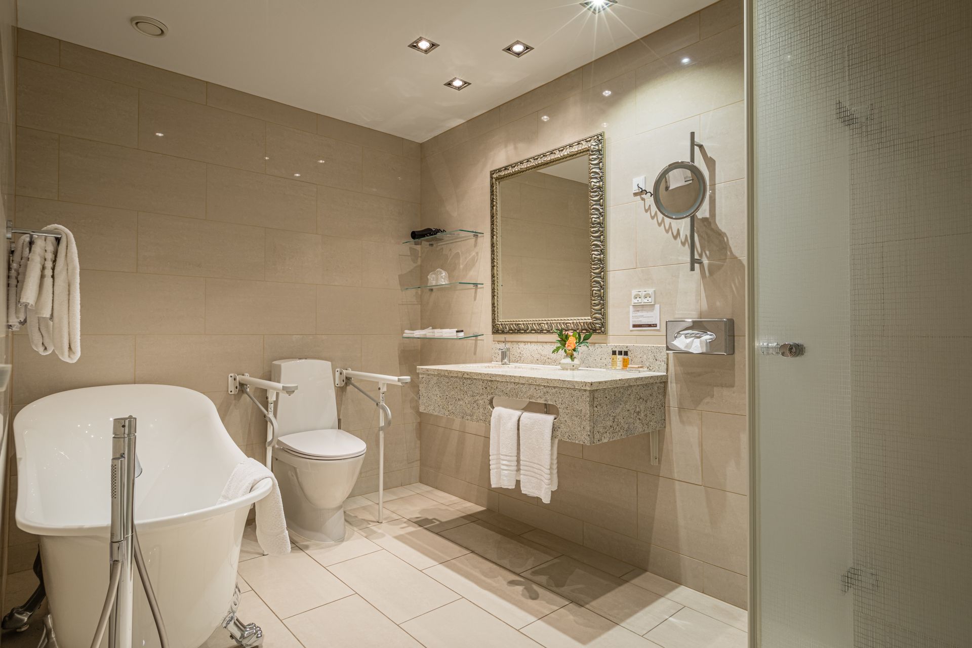 Badeværelse med badekar, toilet og håndvask. Beige fliser på vægge og gulve, handicapvenlige faciliteter.