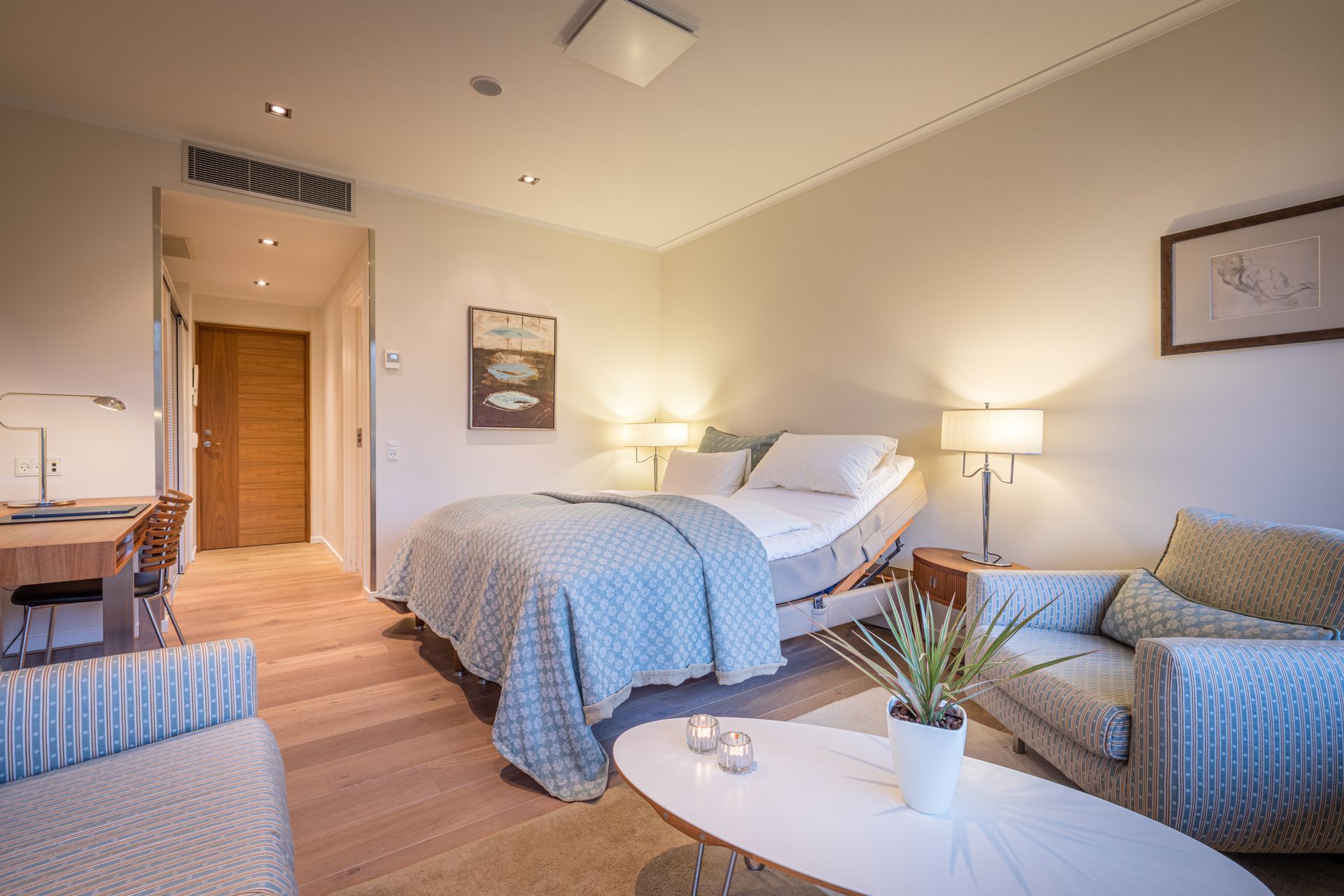 Hyggeligt soveværelse med seng, sofa, skrivebord og sofabord. Trægulv, blød belysning.