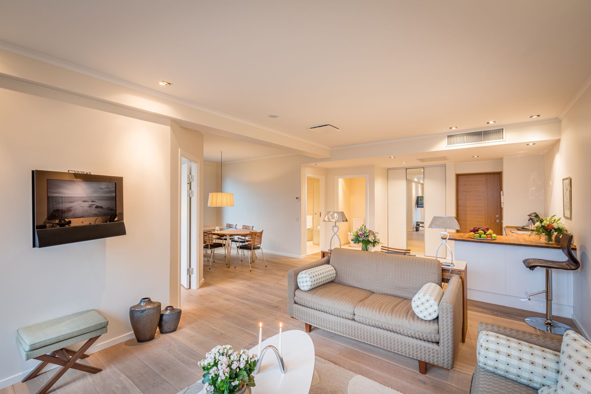 Stue med lyse trægulve, beige sofa og et vægmonteret TV. Spiseplads og køkken i baggrunden.