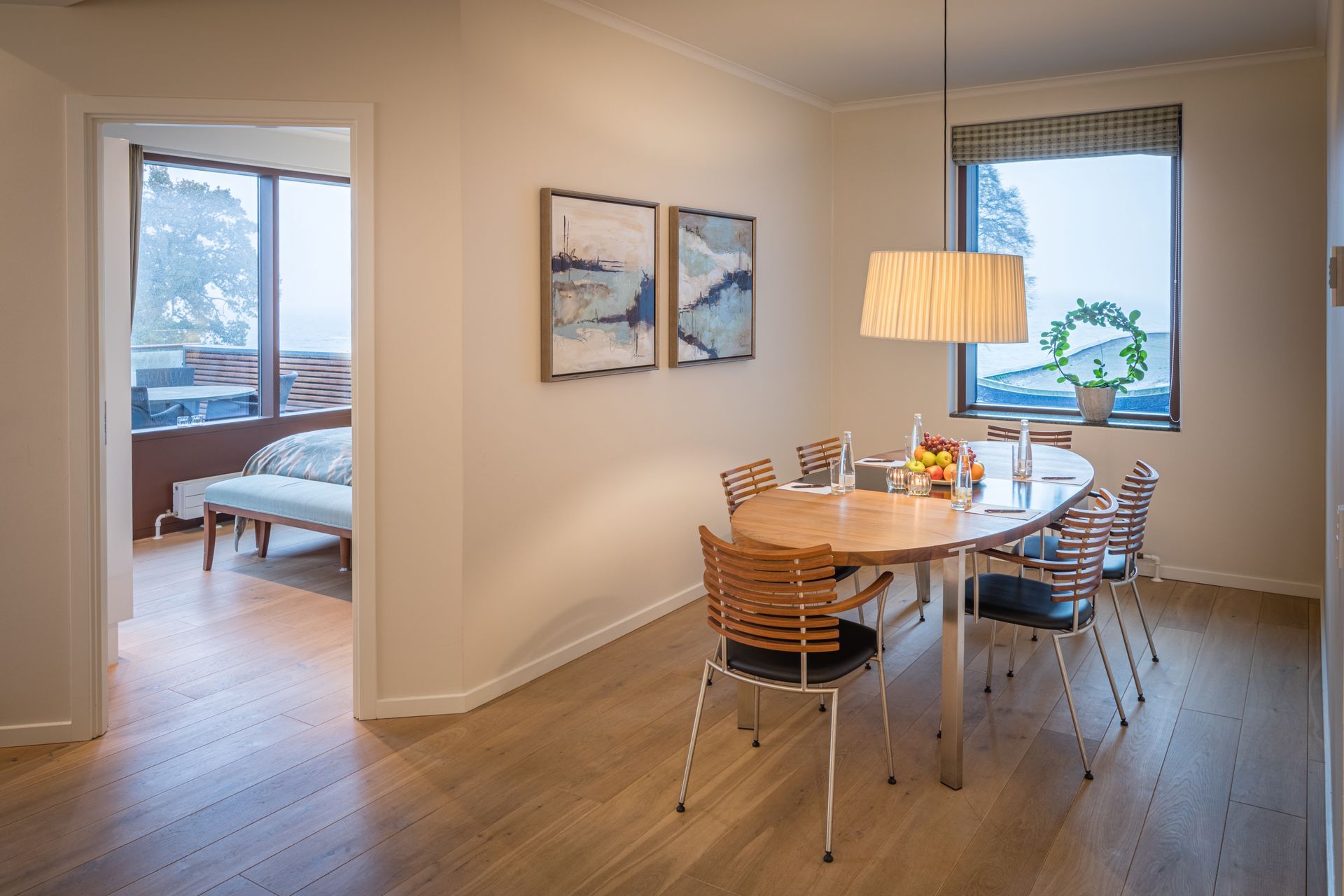 Spisestue med ovalt bord, stole, kunstværker og vindue med udsigt over det fri. En tilstødende døråbning fører til et soveværelse.