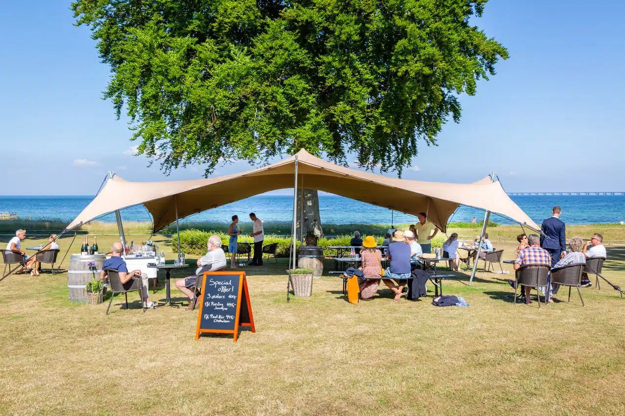 En gruppe mennesker sidder under et telt på en mark nær havet.