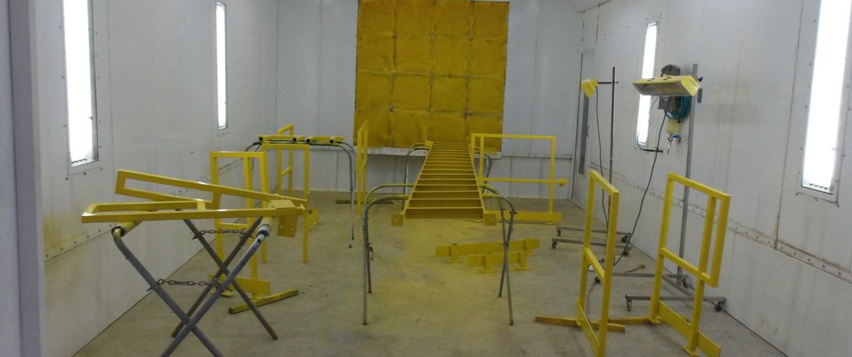 Inside a yellow paint booth, various metal frames are being painted yellow.