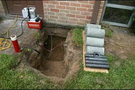 A hole in the ground next to a brick building with a bunch of pipes in it.