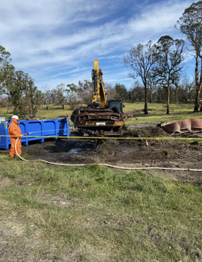 A man in an orange jumpsuit is standing next to a yellow excavator in a field.