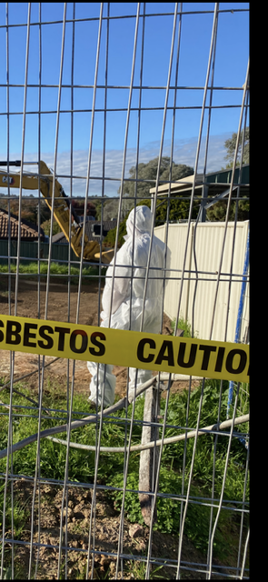 A person in a protective suit is standing behind a fence with asbestos caution tape.