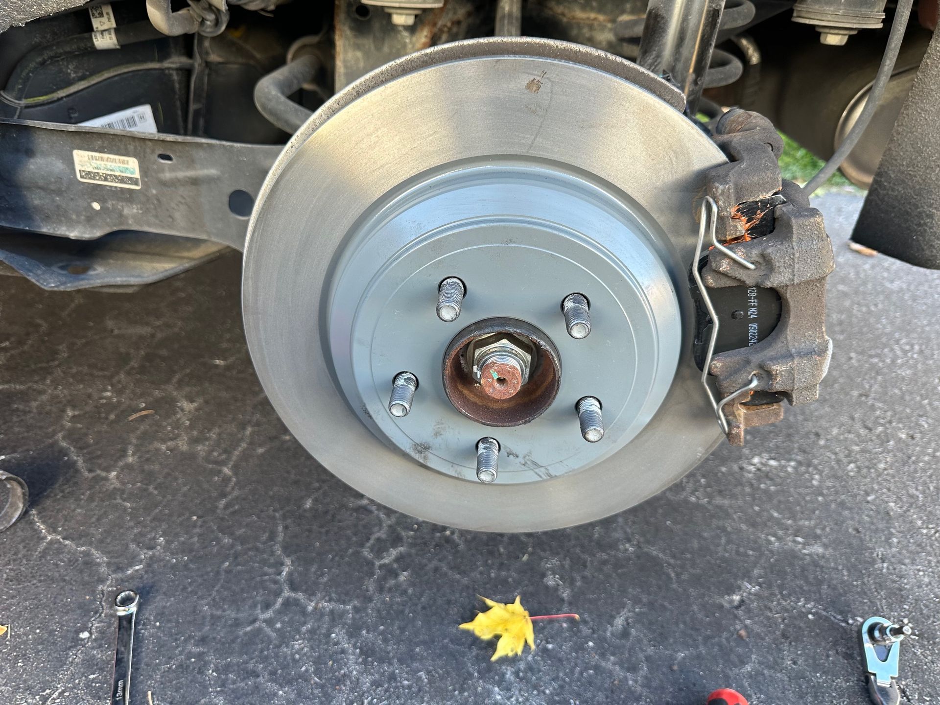 Car's rear wheel with the brake rotor and caliper exposed during repair. Gray metal, bolts visible, on asphalt | Campbell’s Fix All LTD