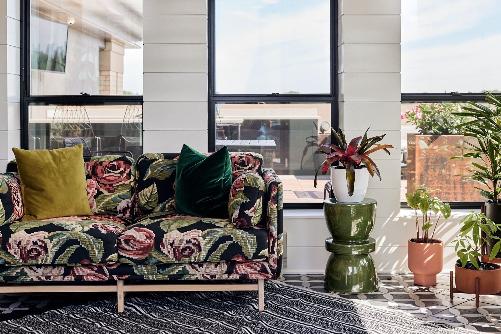 Floral sofa with pillows sits by windows and plants.