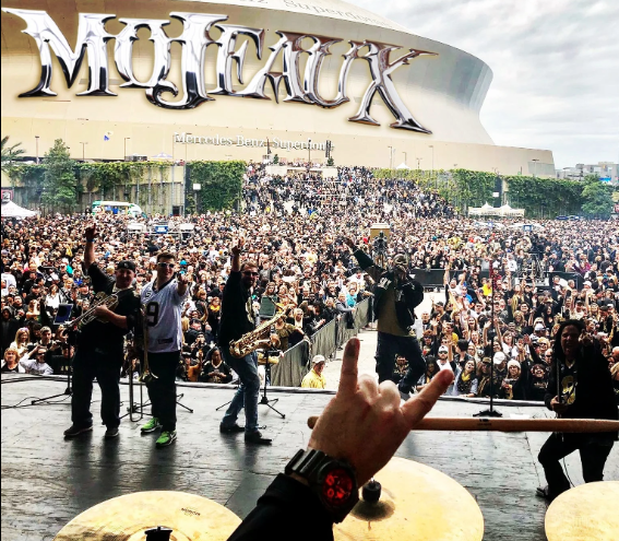 Concert crowd and performers on stage in front of a large “Mojaxx” sign, viewed from behind a drum set