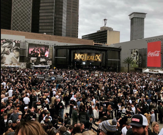 Crowd at an outdoor concert with a large stage and city buildings in the background