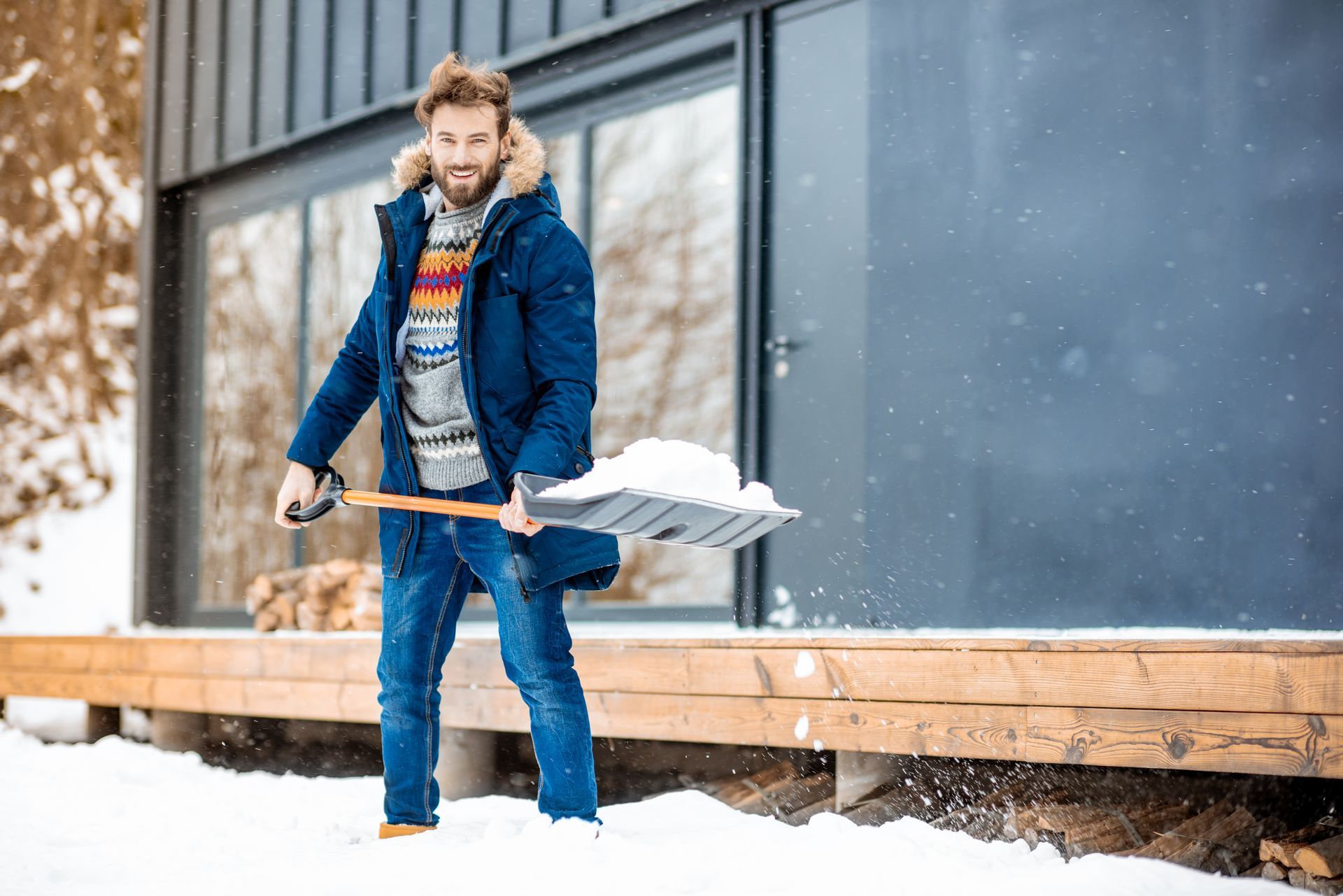Man shoveling snow from a wooden porch wearing a blue parka, jeans, and a patterned sweater.