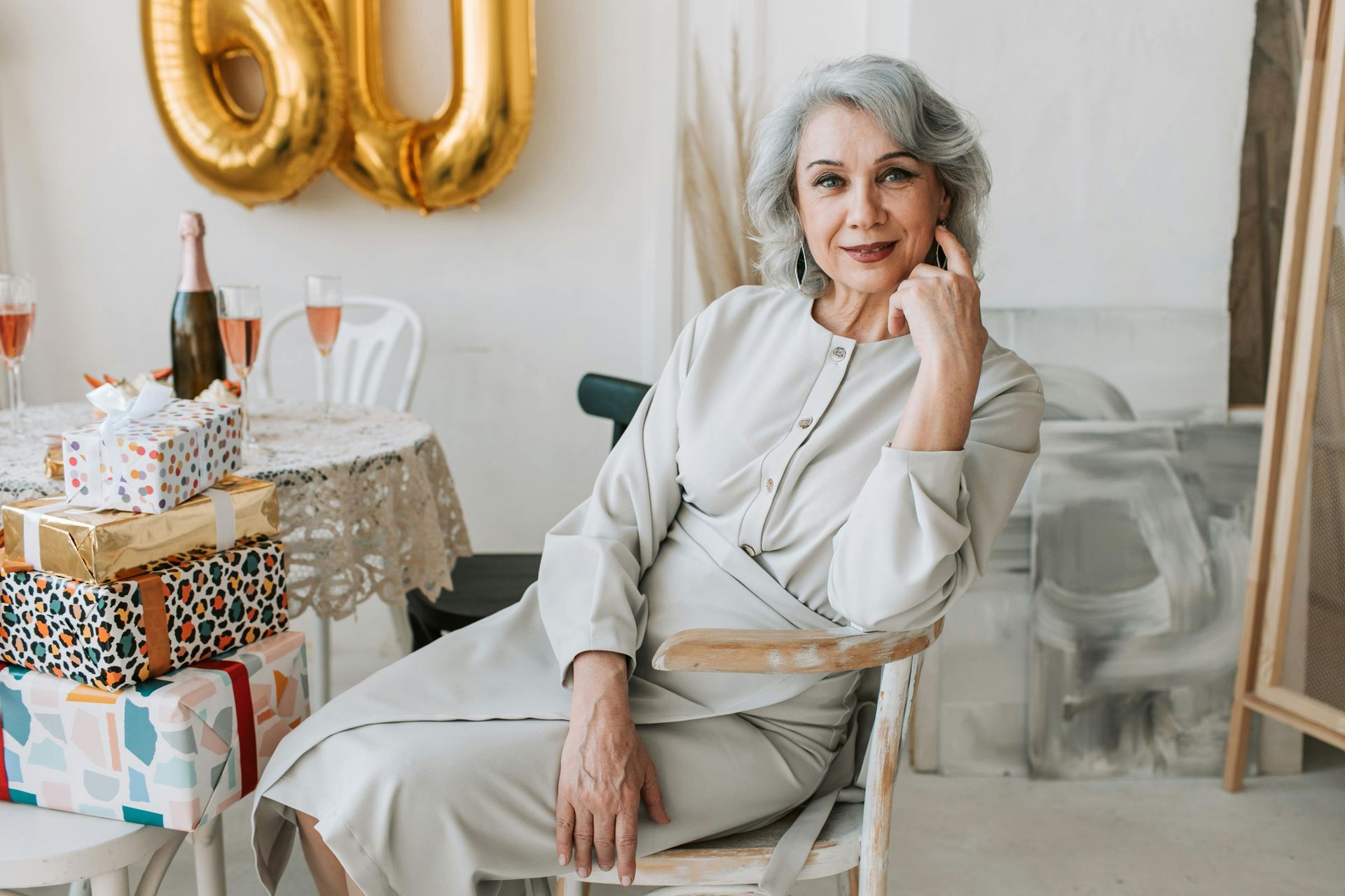 Woman with gray hair seated at a table, celebrating a 60th birthday. Presents and balloons in background.