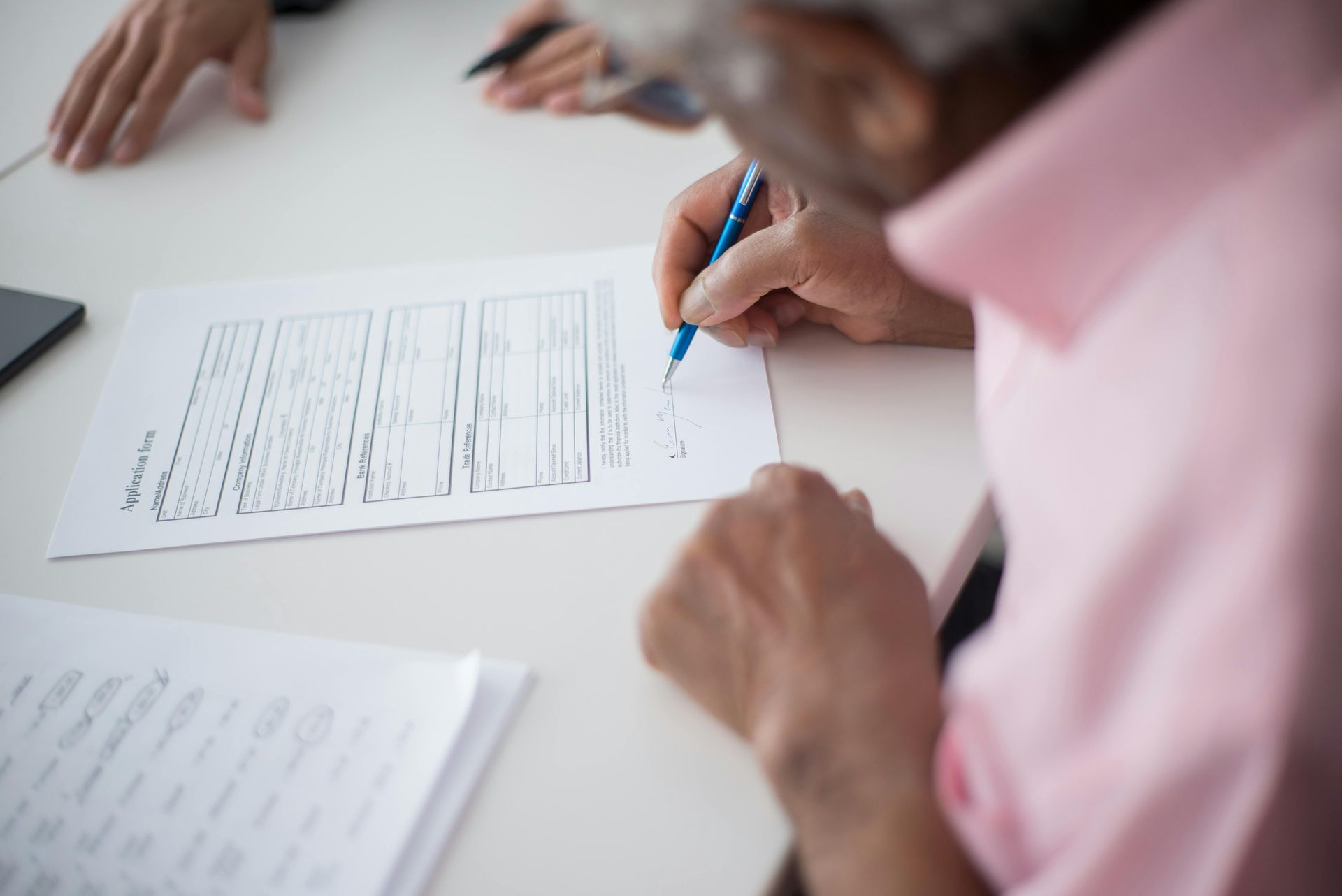 An older person in a pink shirt signs a document at a white table. Others' hands are visible nearby.