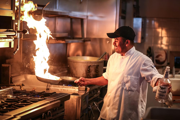 Chef in white coat cooking at a restaurant stove with a large flame rising from the pan
