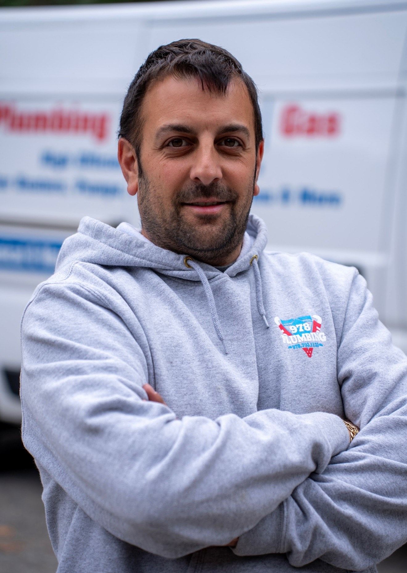 A man in a gray hoodie is standing with his arms crossed in front of a white van.