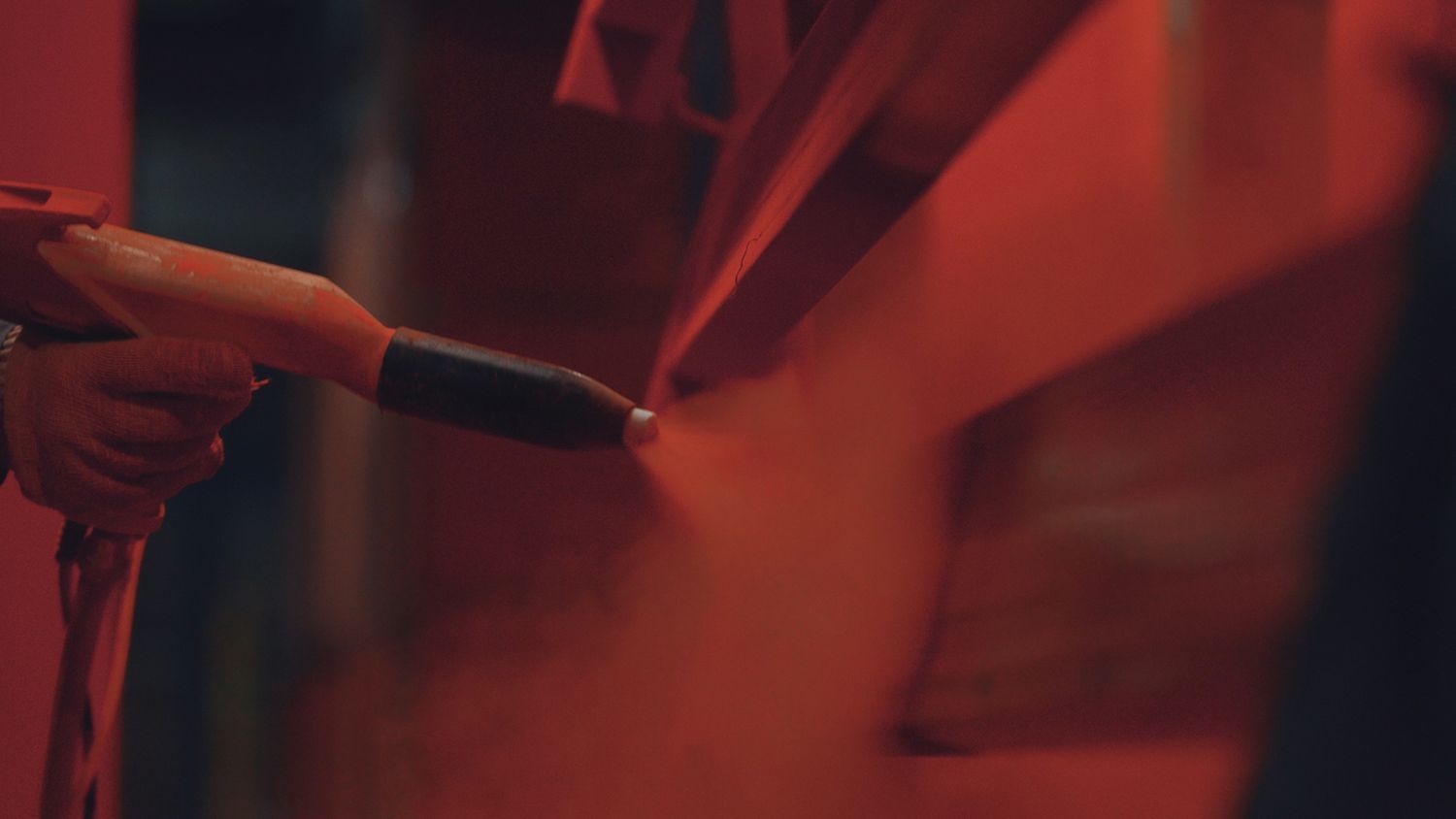 View of a worker's hand with gloves and a spray gun applying red powder coats to a metal surface.