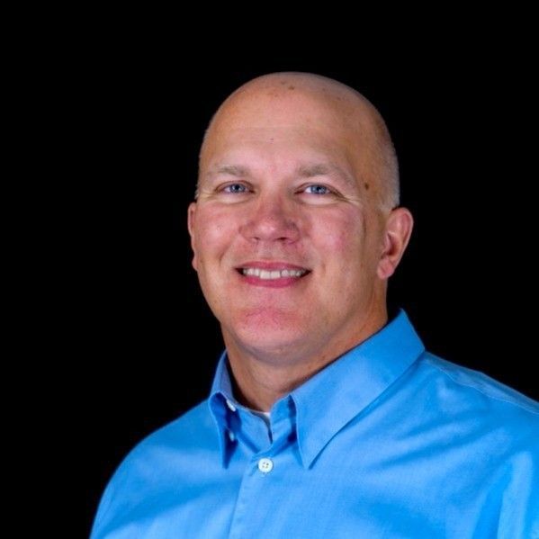 Bill Magnuson. Bald man with blue shirt smiles against a black background.