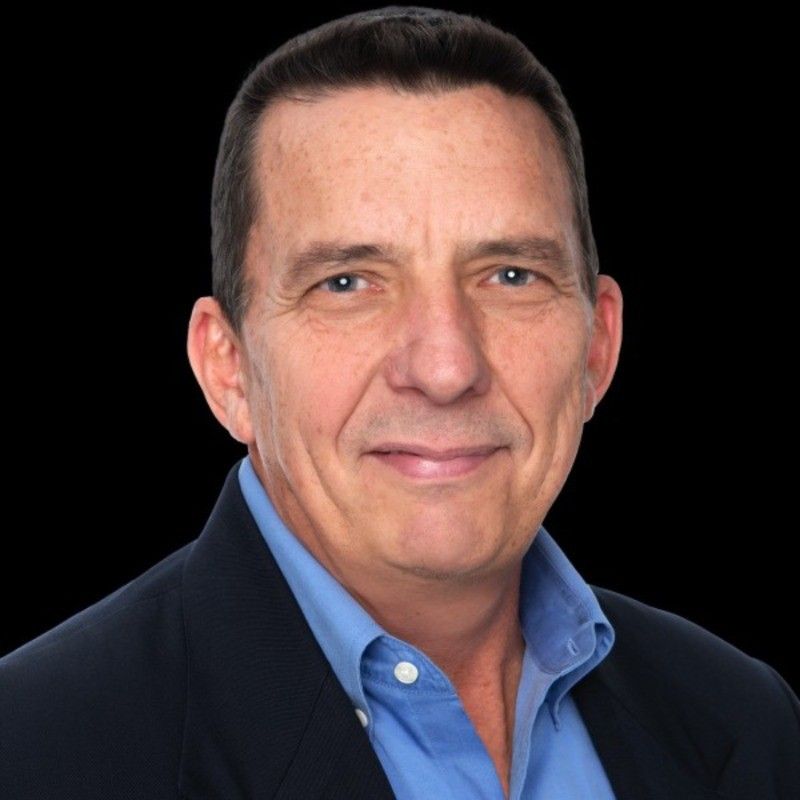 Dan Keck. Man with short dark hair in a blue shirt and navy jacket smiles. Black background.