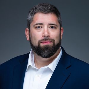 Eli Coulton. A man with a beard in a suit jacket poses for a headshot.