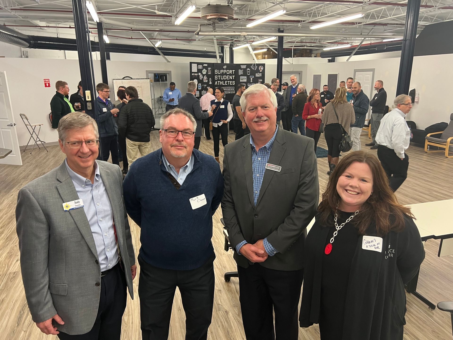 Four people pose in front of a gathering. One in black dress, others in business attire, smiling. Open room, wood floor.