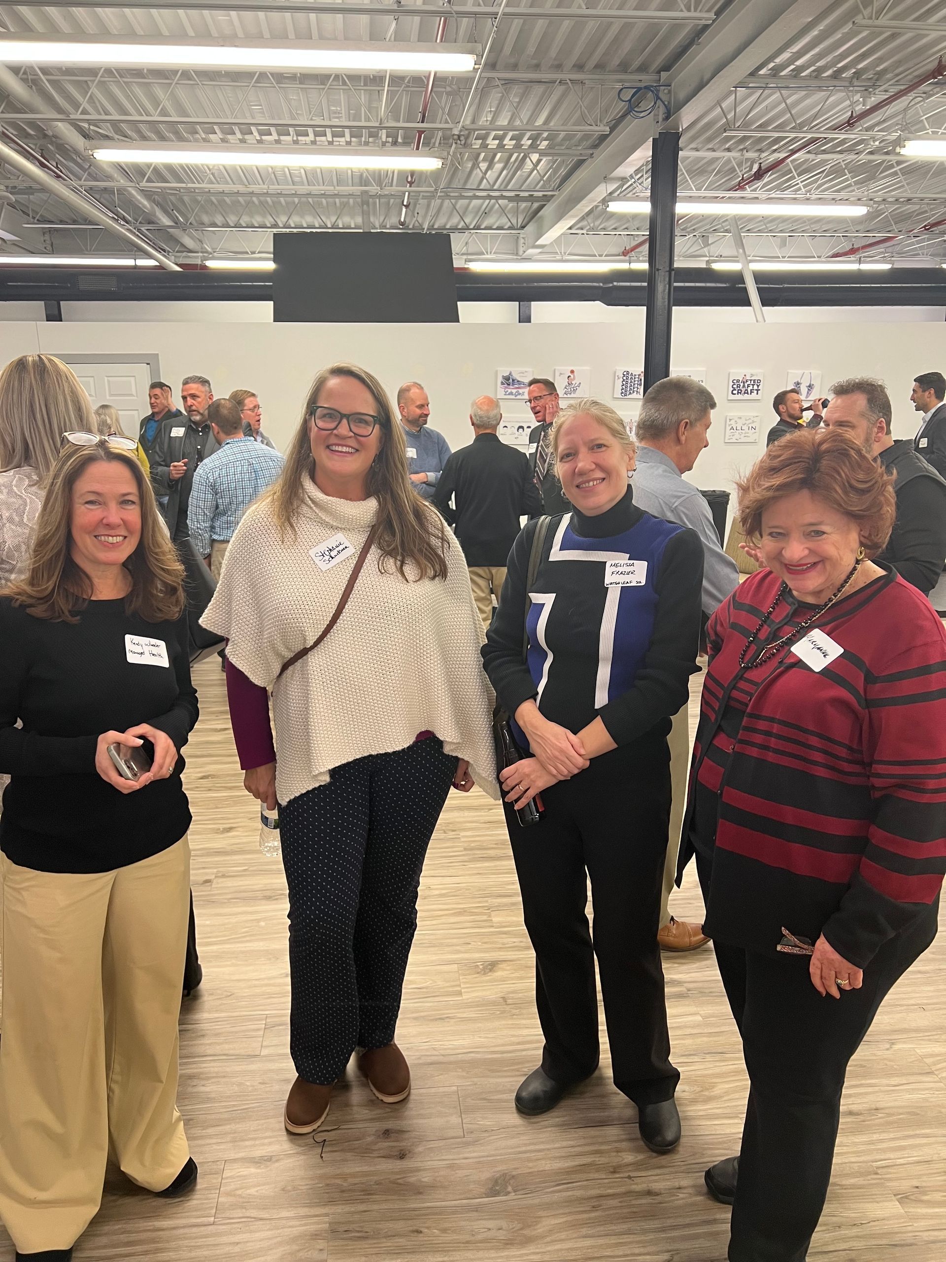 Four women at an event, smiling. One in white sweater, another in black, one in red sweater, and one in tan pants. Others in background.