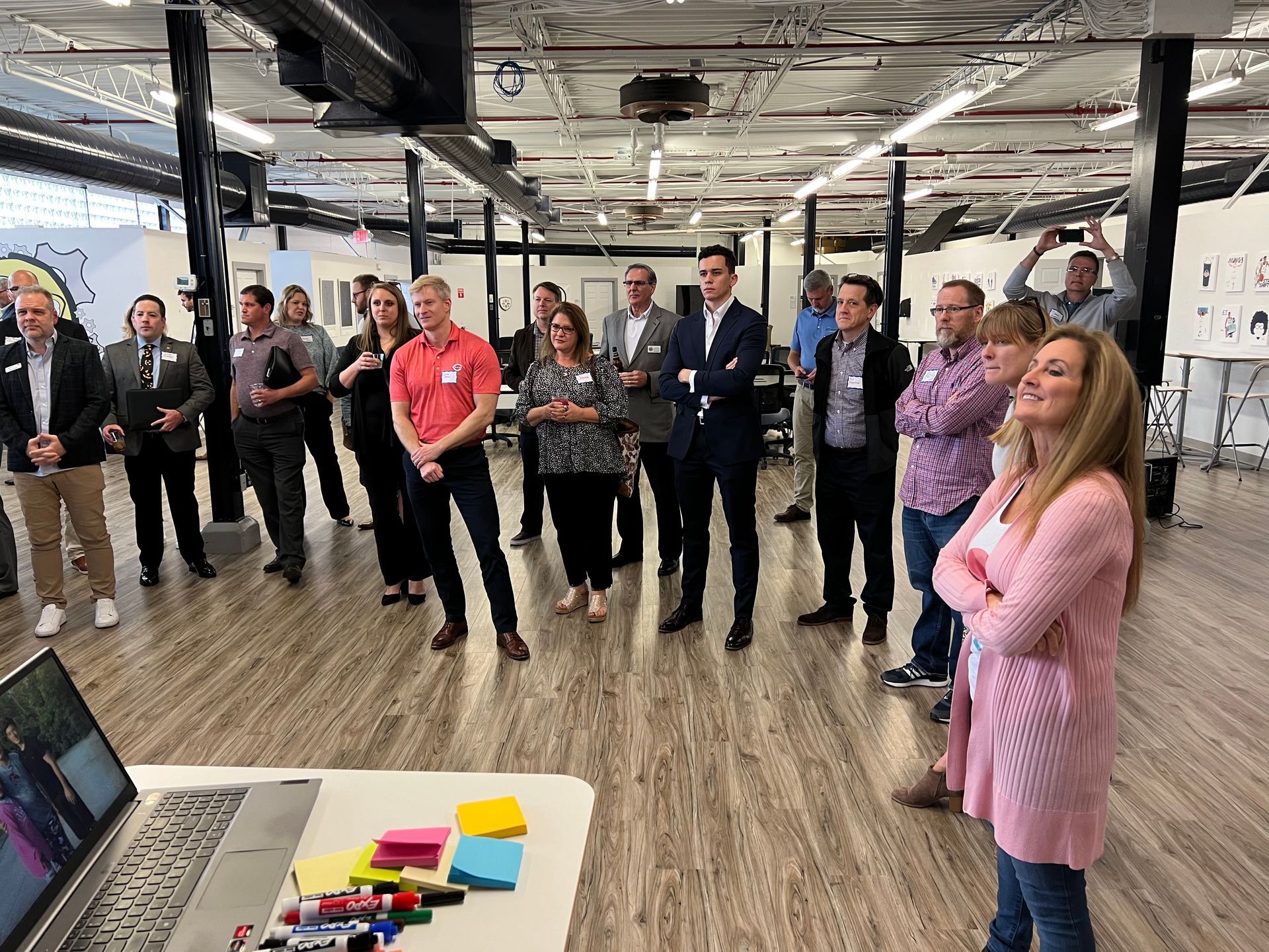 Group of people in a large room, watching a laptop on a table; some are smiling.