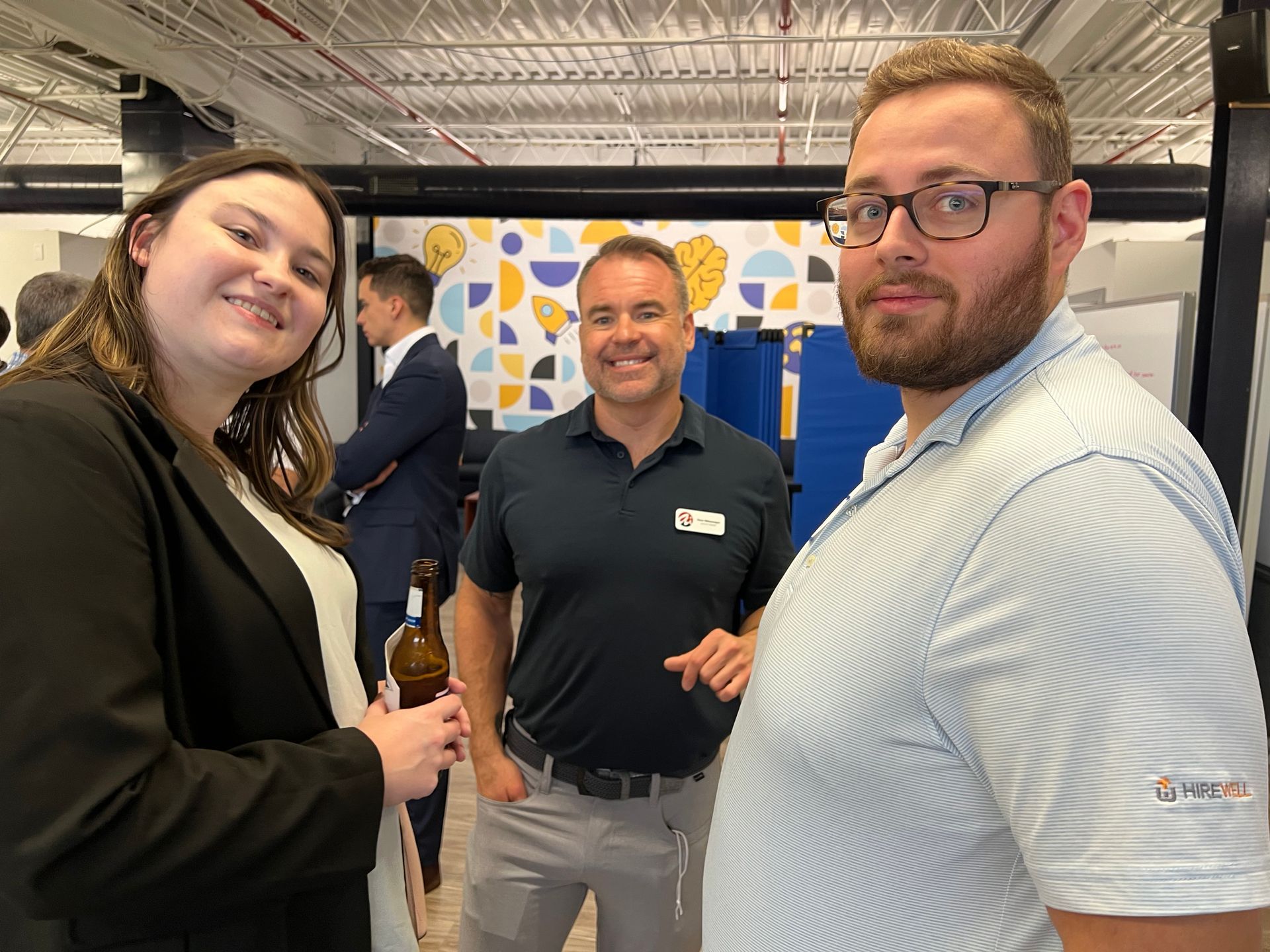 Three people smiling for a photo. A man in the center wears a polo, while the others are dressed in business attire.