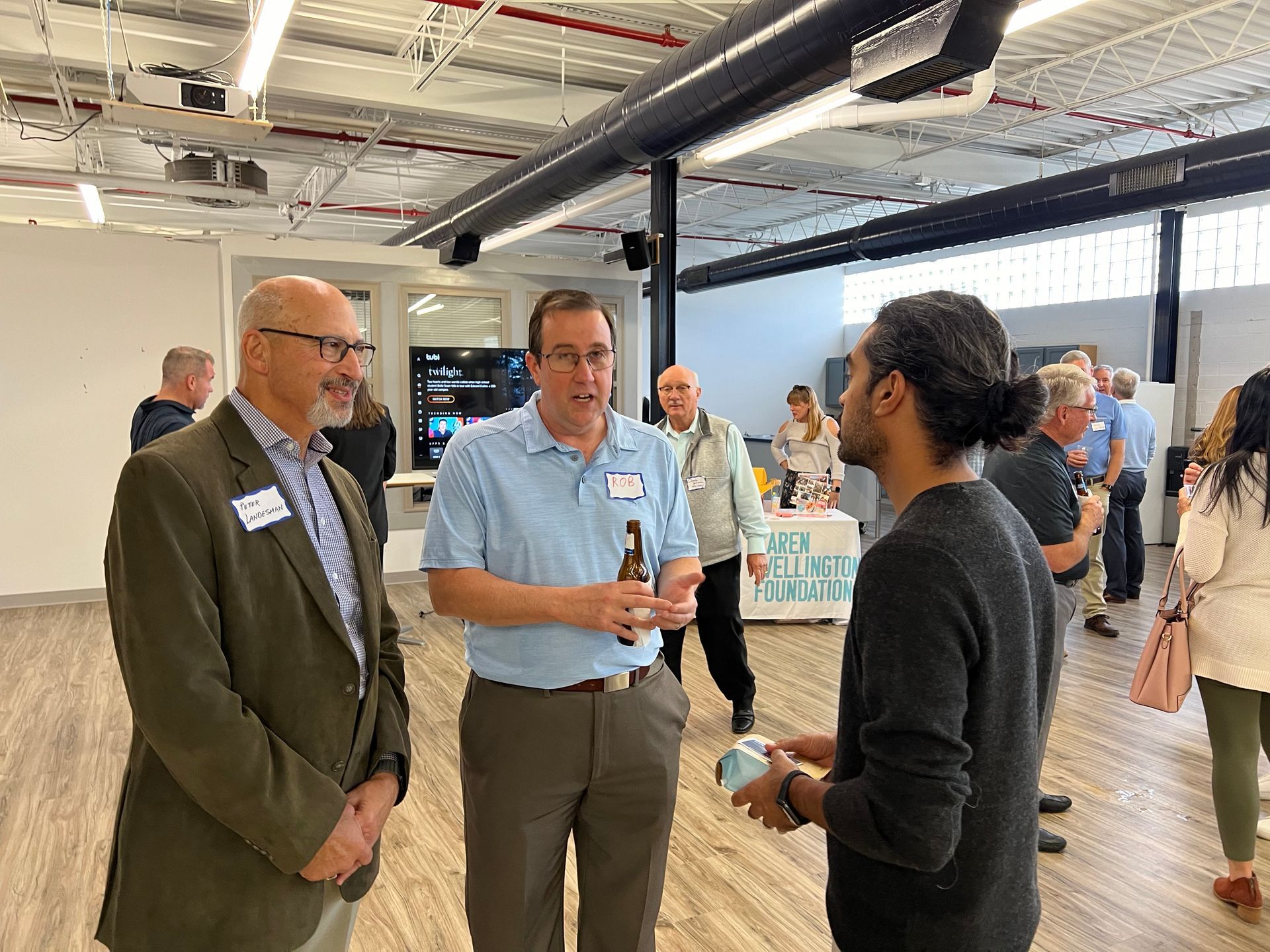 Three men conversing at an event, one holding a drink. Other attendees are in the background.