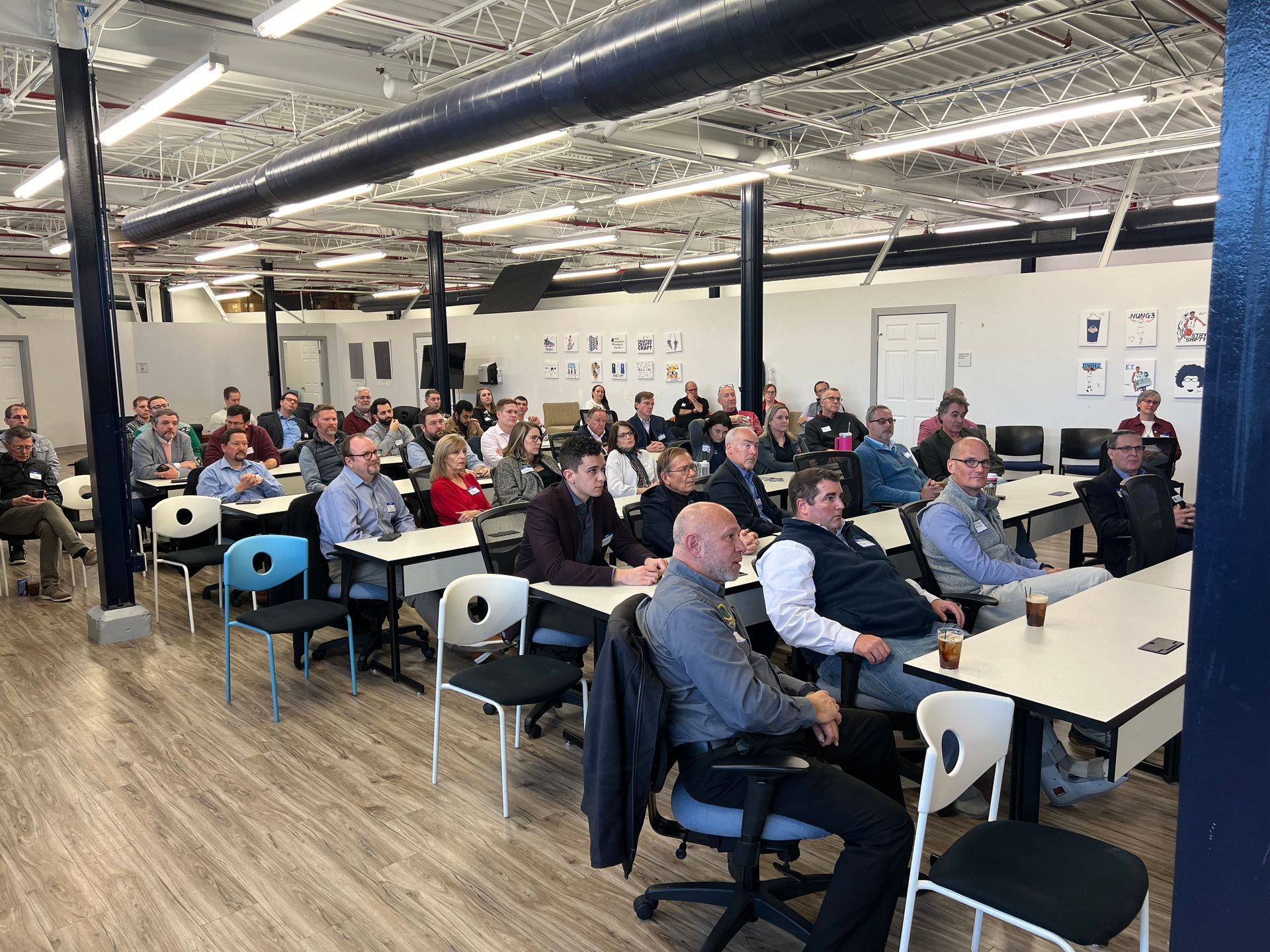 People in a presentation setting. Attendees seated at tables, a few looking at the presenter. Bright room.