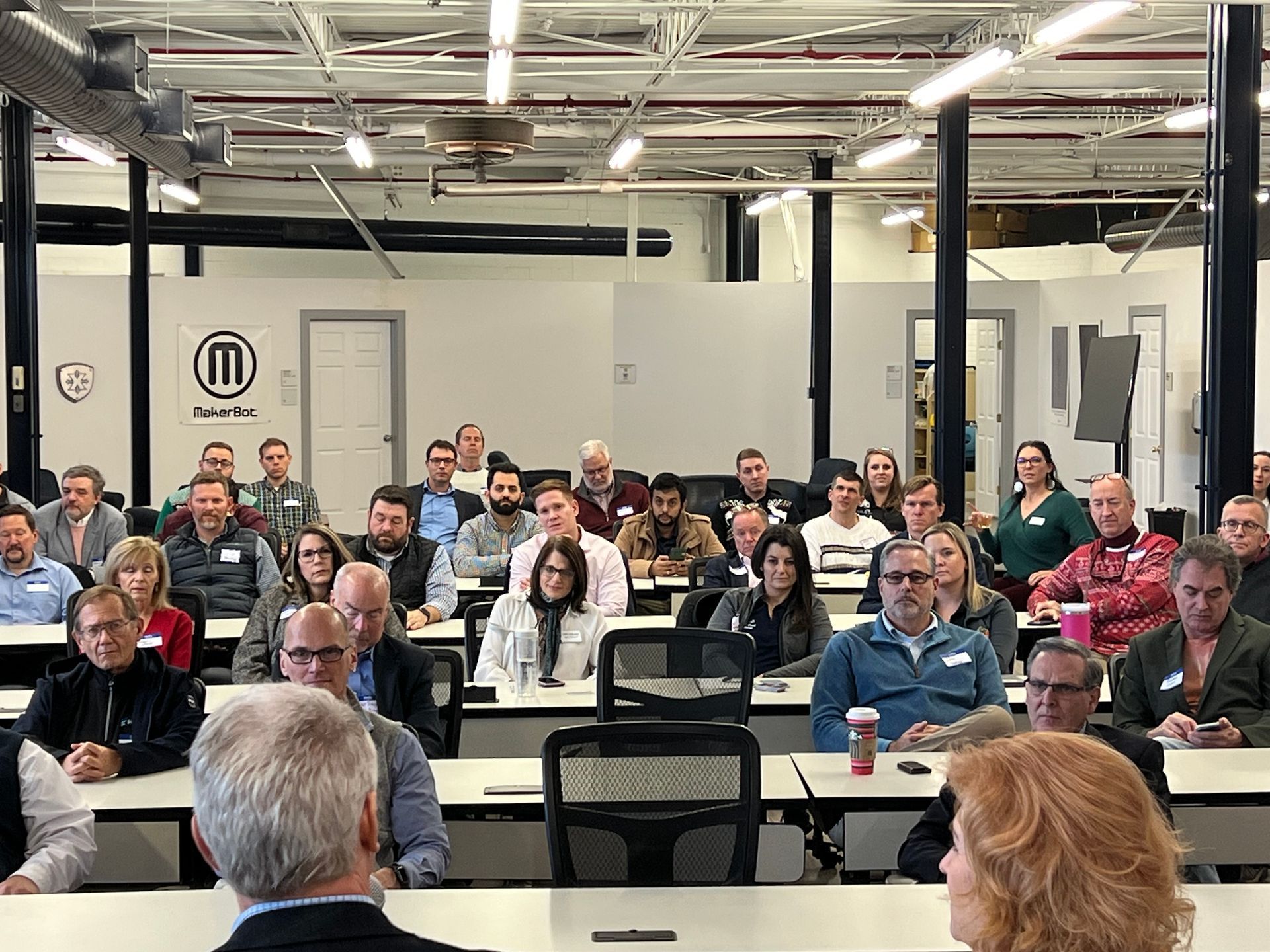 Audience in a classroom setting, listening to a speaker. White walls, rows of tables, and overhead lighting.