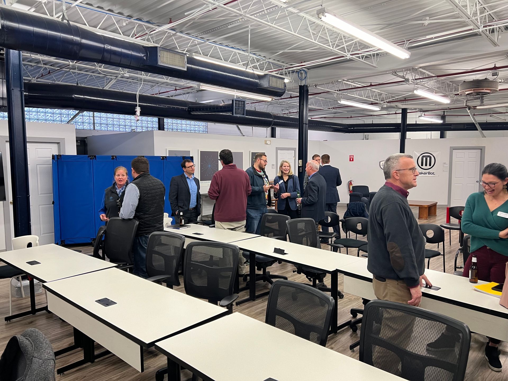 People gather in a large room with long tables and chairs.  Some are talking in small groups. Exposed ceiling, blue panels.
