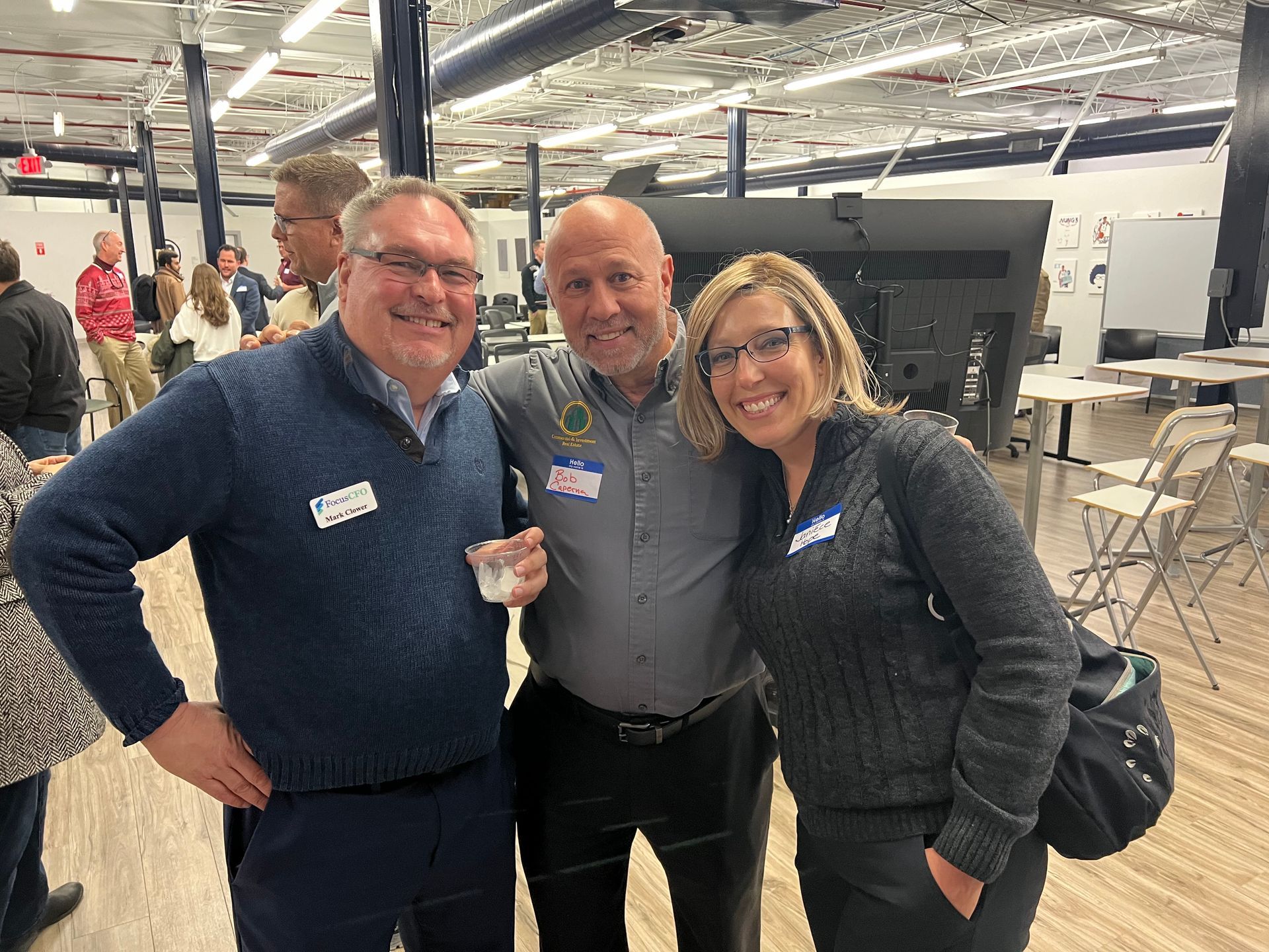 Three people smiling and posing together indoors; one holds a drink.