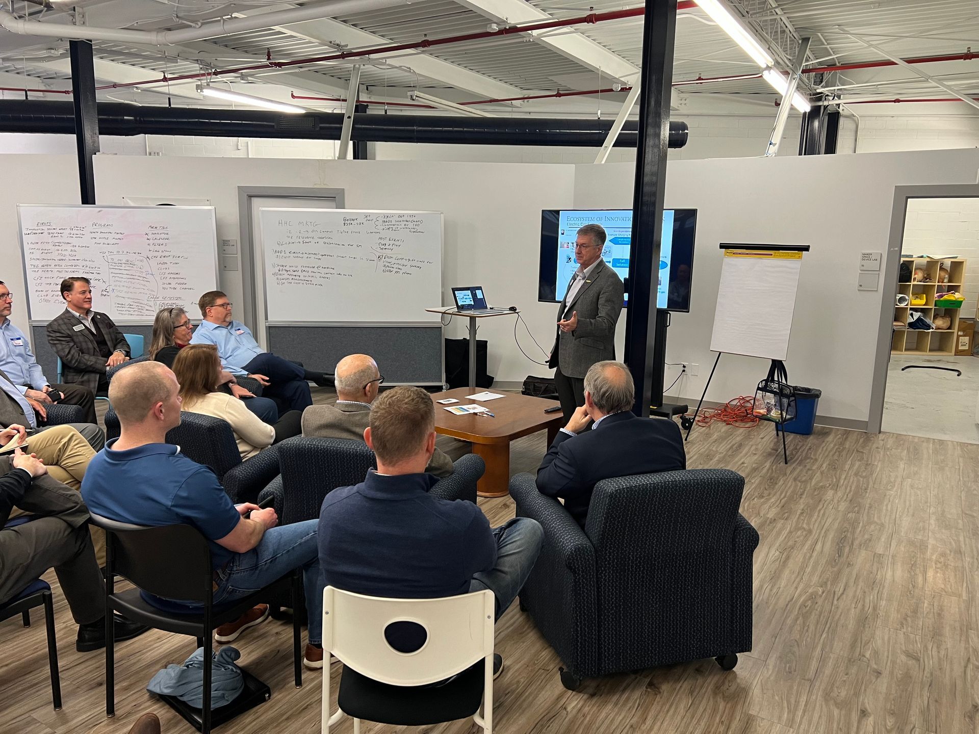 Man giving presentation to a seated audience in a bright room. A screen and whiteboards are visible.