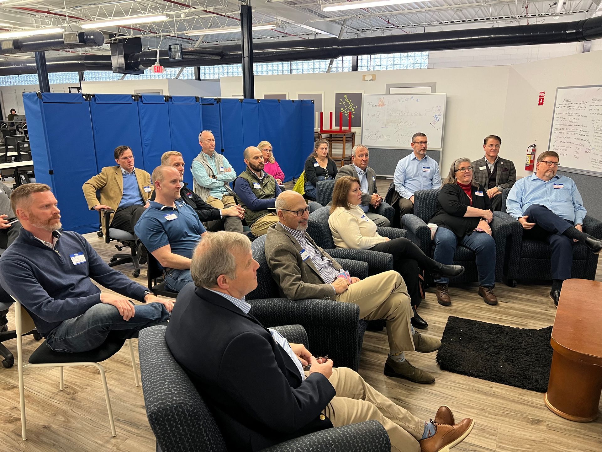 A group of people seated and listening in an office setting. Some are smiling, some are taking notes.