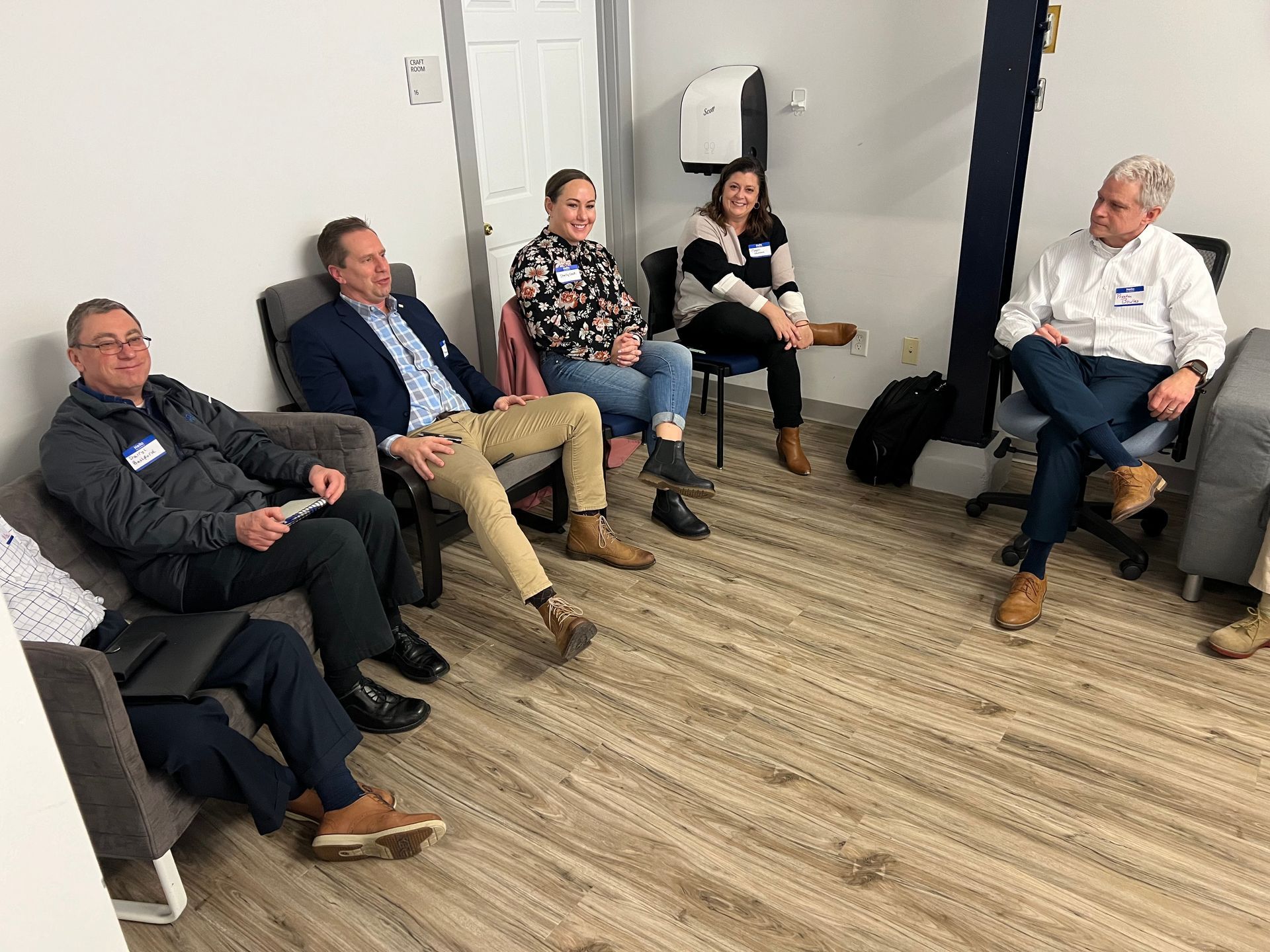 Five people seated in a circle, engaged in conversation. Gray and neutral colored room, carpet, and chairs.