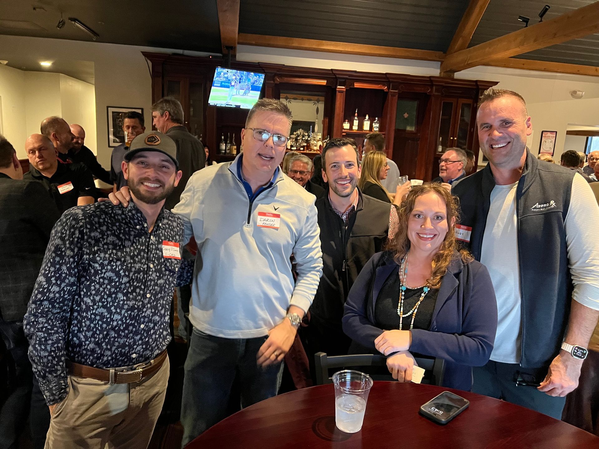 Group of people at a bar, smiling. Men and women in casual attire. Dark bar setting.