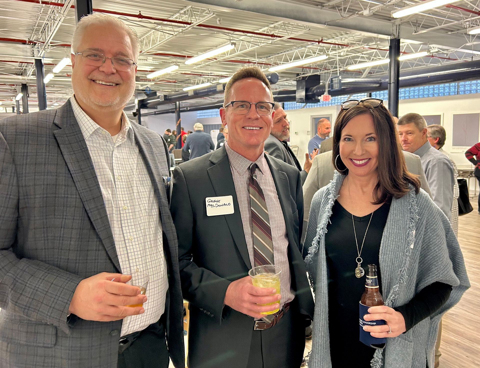 Three people at an event: two men and a woman, smiling and holding drinks, indoors.