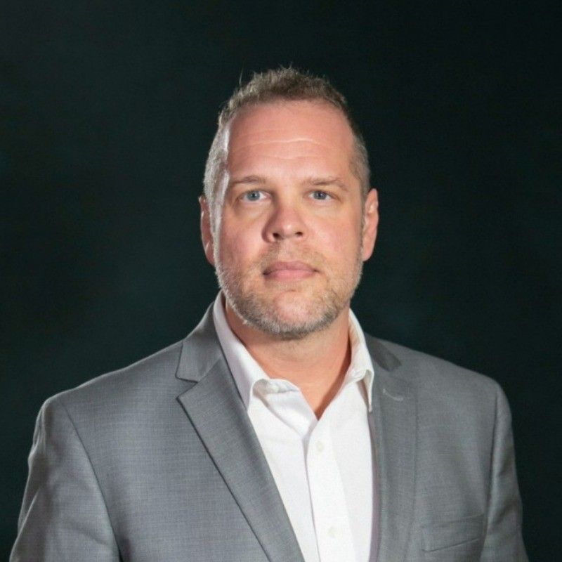 James Sayer. Man in gray suit and white shirt, looking at the camera. Dark background.