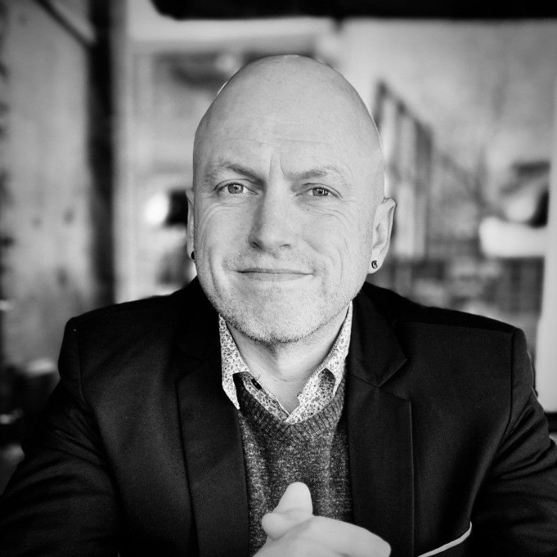 Jon Harney. Bald man with a slight smile, wearing a suit jacket, sweater, and patterned shirt. Indoors.