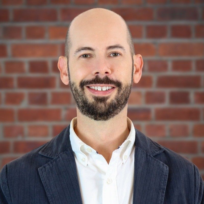 Joshua VanDixhorn. Bald man with a beard smiling, wearing a white shirt and blue blazer, standing in front of a brick wall.
