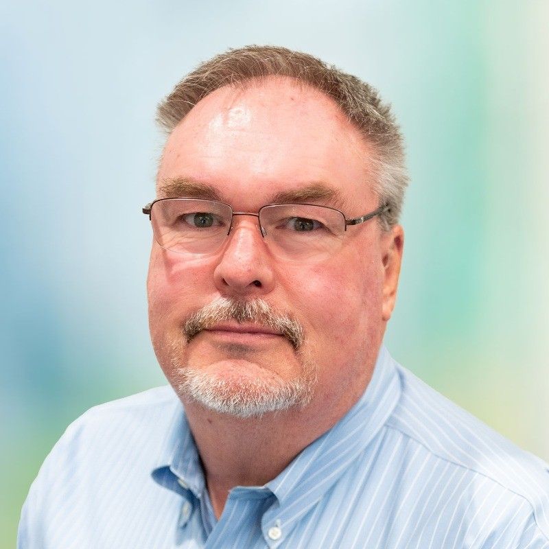 Mark Clower. Man with glasses and a short beard, wearing a blue shirt, looks at the camera.