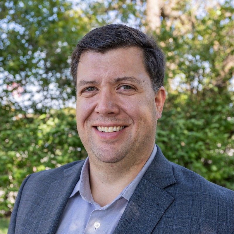 Mark Mansur. Man in a gray blazer and blue shirt smiles broadly outdoors, trees in the background.